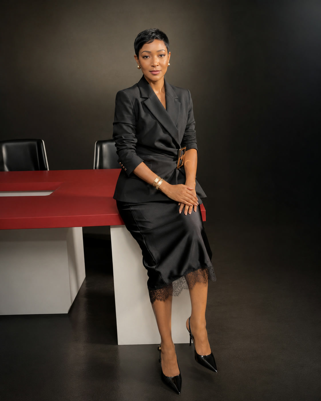 Yolanda Robert in a black tailored outfit, seated at the edge of a red studio desk — editorial service portrait